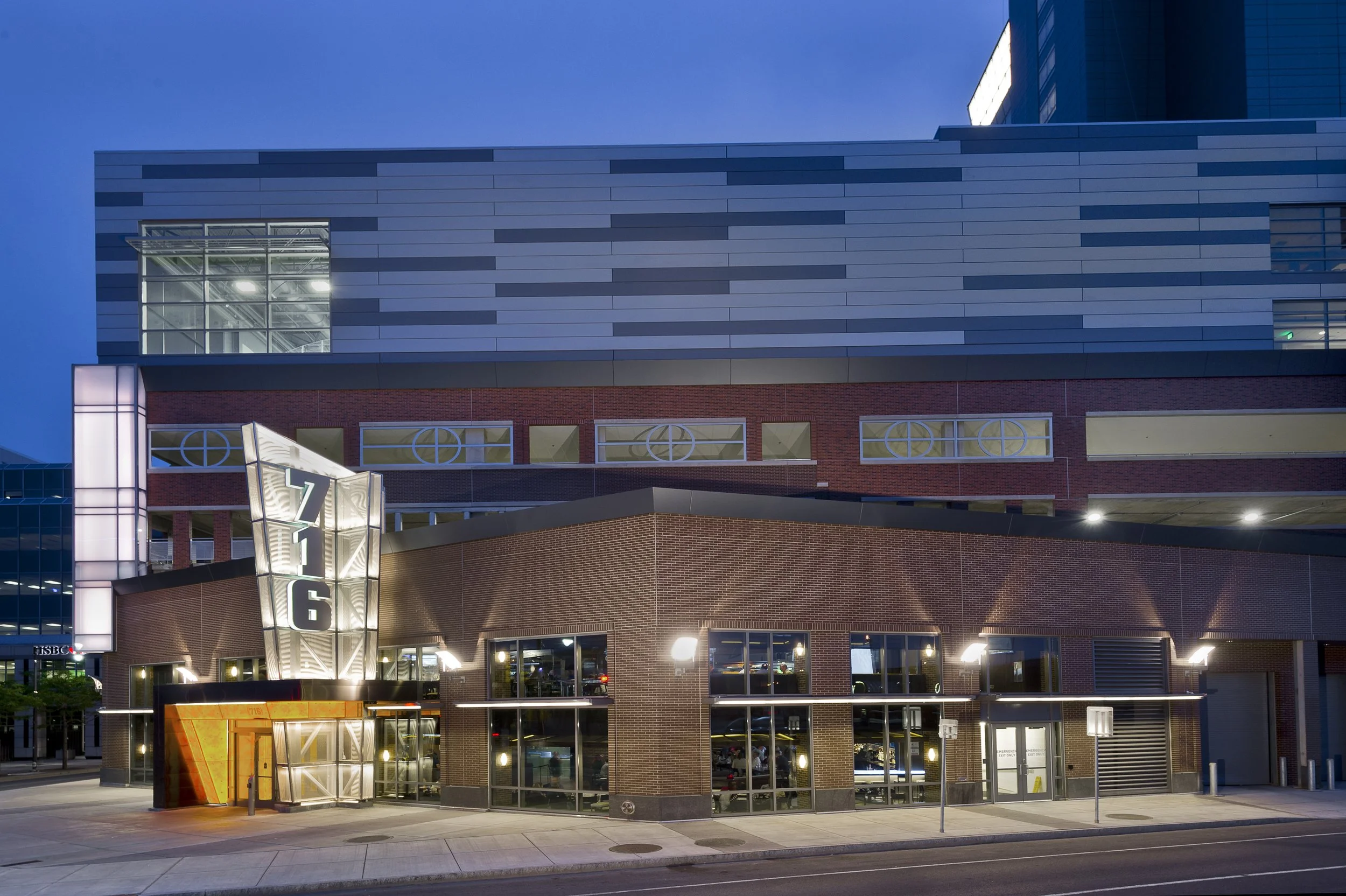 A modern multi-story building with brick and metal exterior, large windows, a prominent lit sign with the number 7 and 6, and entrance area illuminated at night.