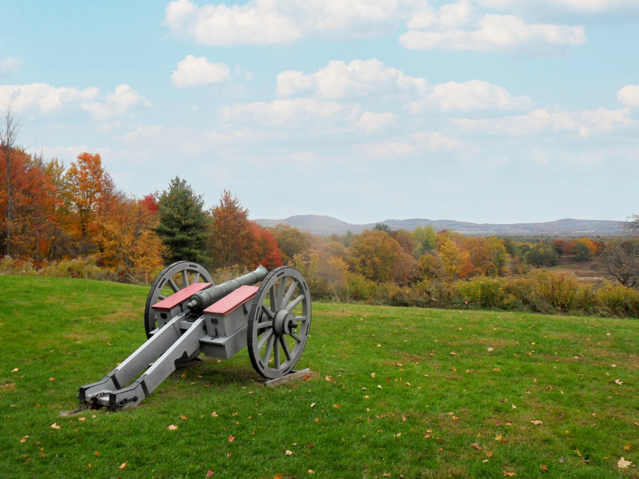 An outdoor scene with a cannon on green grass, colorful fall trees in the background, rolling hills in the distance, and a clear sky with clouds.