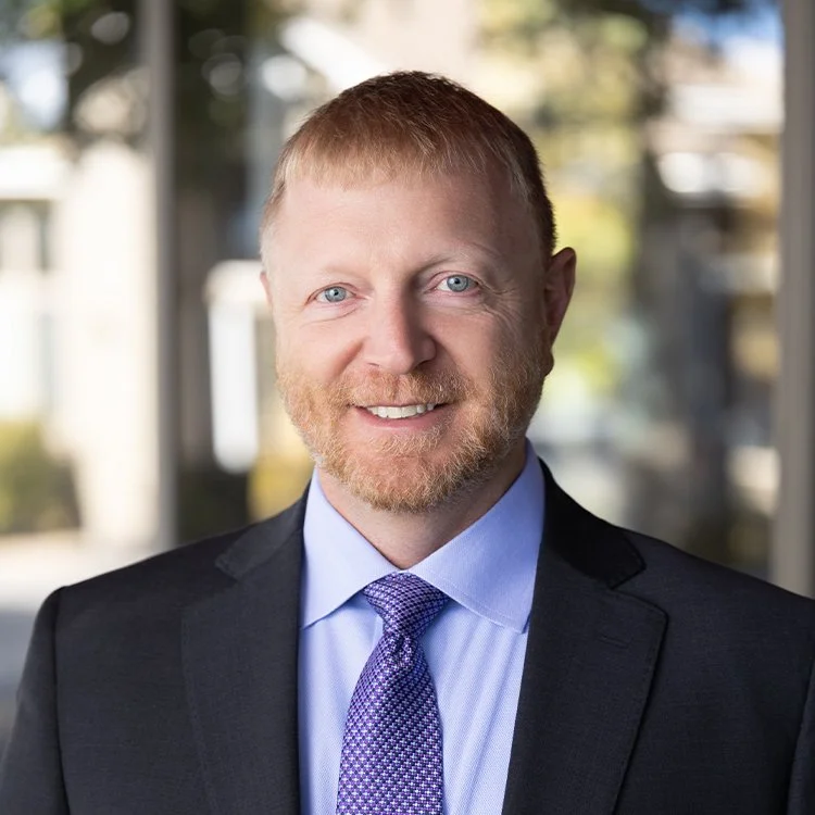 A man with light skin, red hair, and a beard smiling, wearing a black suit, light blue shirt, and purple tie, standing indoors with large windows and blurred background.
