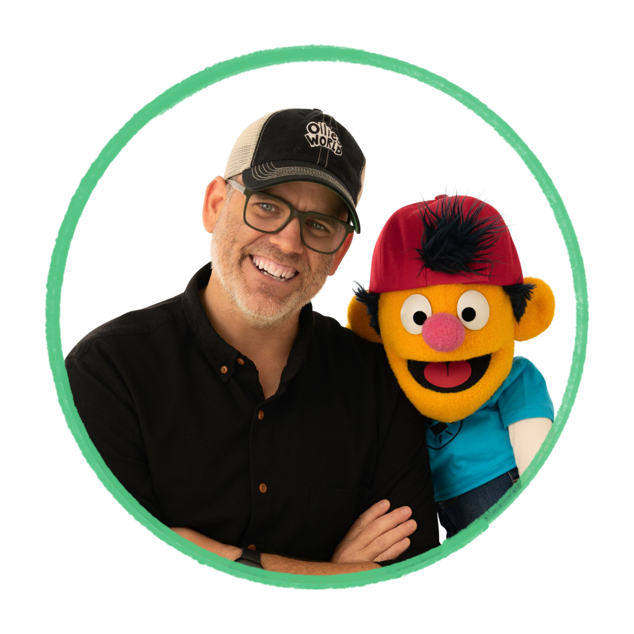 A smiling man with glasses and a black cap standing next to a puppet character with a red cap, yellow face, and colorful attire.