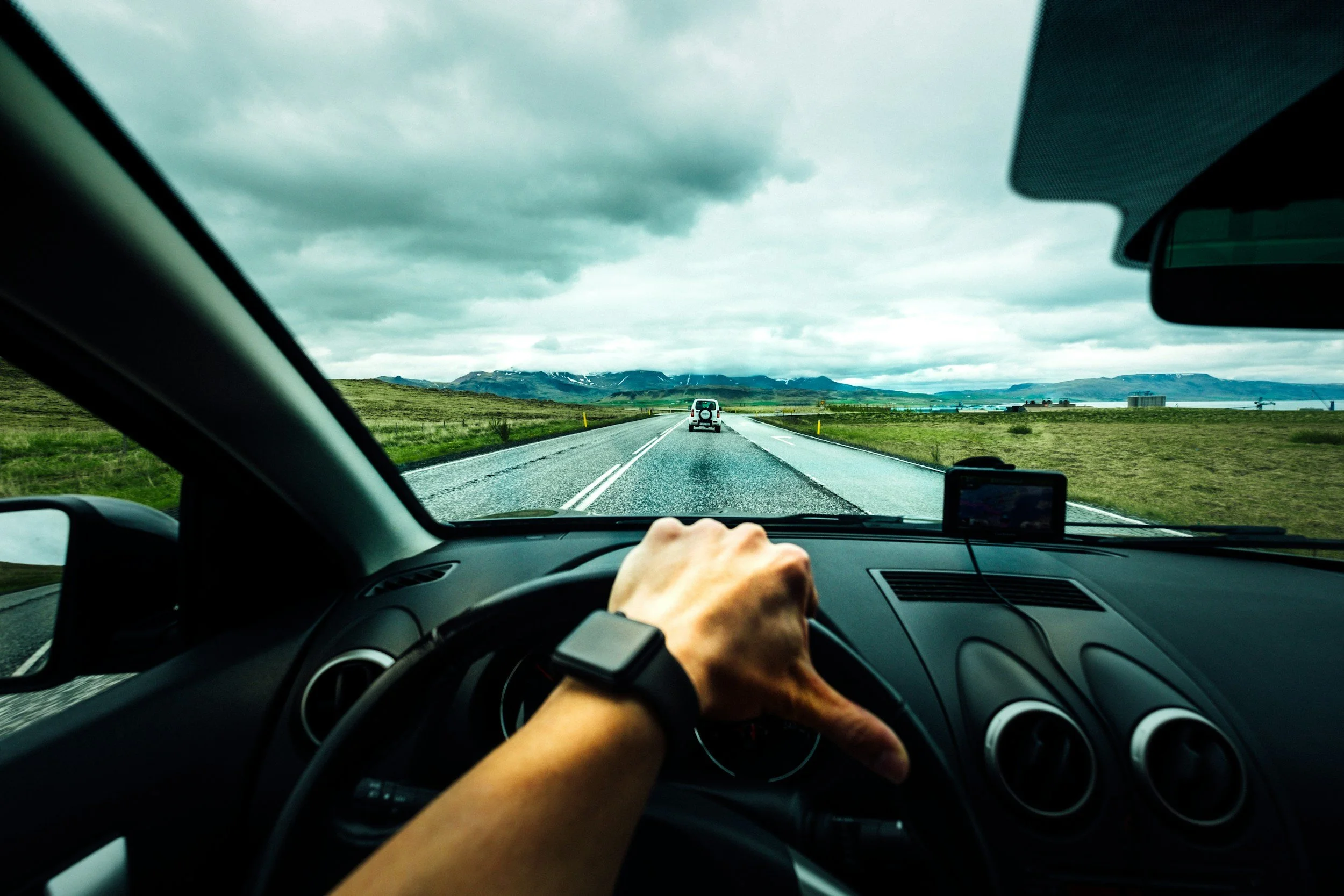 Inside a car on a highway surrounded by open landscape and mountains, with a driver’s hand visible on the steering wheel and a road trip scene.