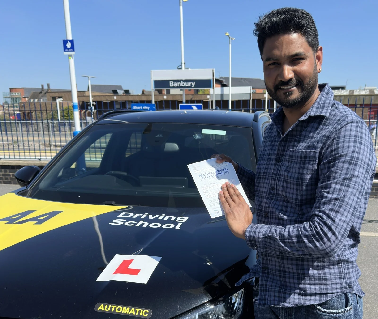 A man with dark hair and a beard holding a practical driving test pass certificate stands next to a black driving school car with a yellow and black 'AUTOMATIC' sticker and a learner 'L' plate, smiling at the camera outside a train station with a 'Ba