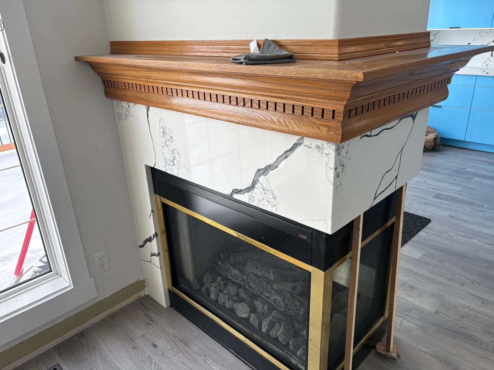 A fireplace with a white marble surround with gray veining and a black and gold metal frame, topped with a wooden mantel shelf, in a modern living space with light wood flooring and a sliding glass door.