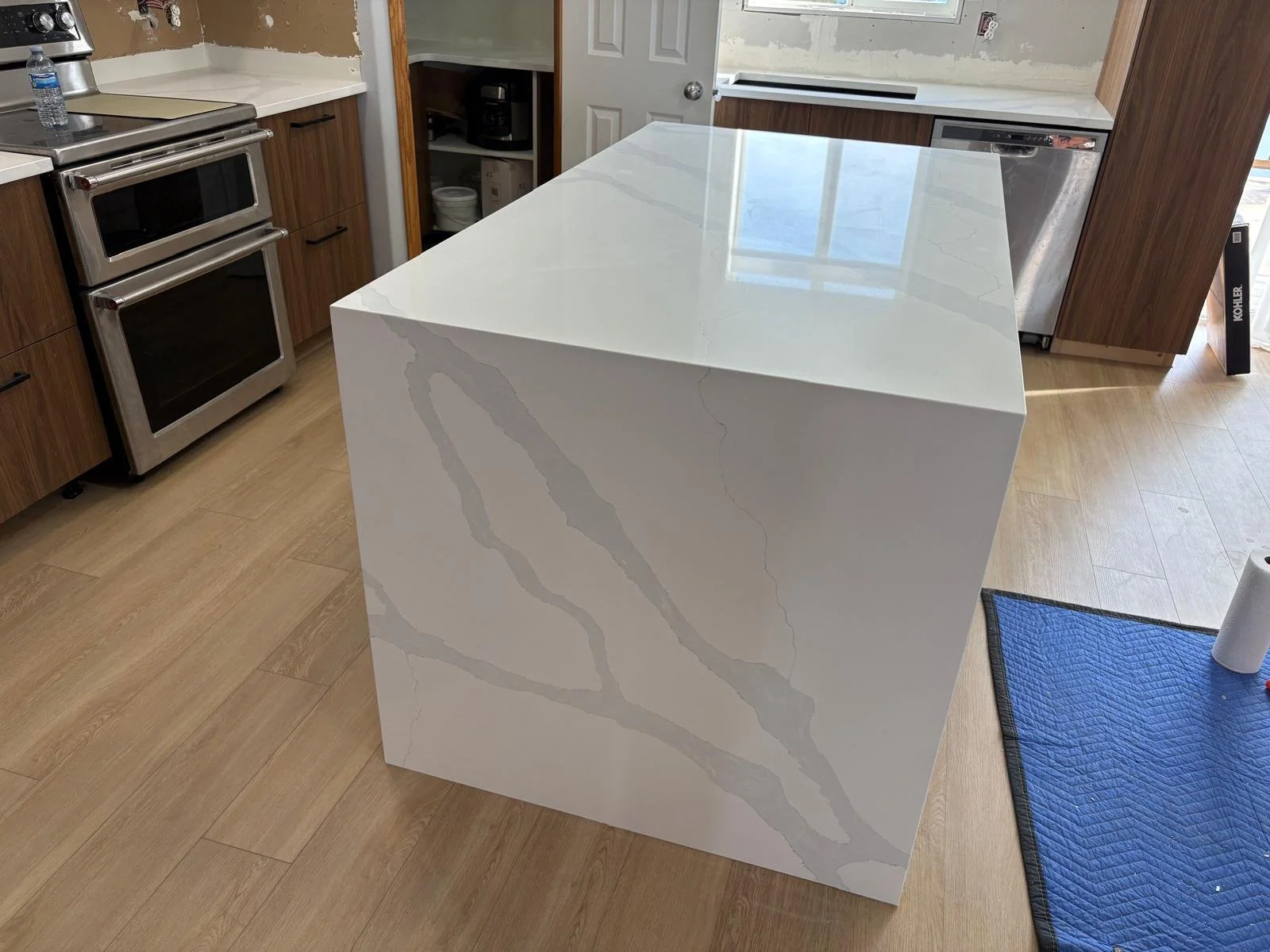 A kitchen island with a white marble countertop featuring gray veining, situated in a kitchen with wooden cabinets, a stove, and a small open cabinet area.