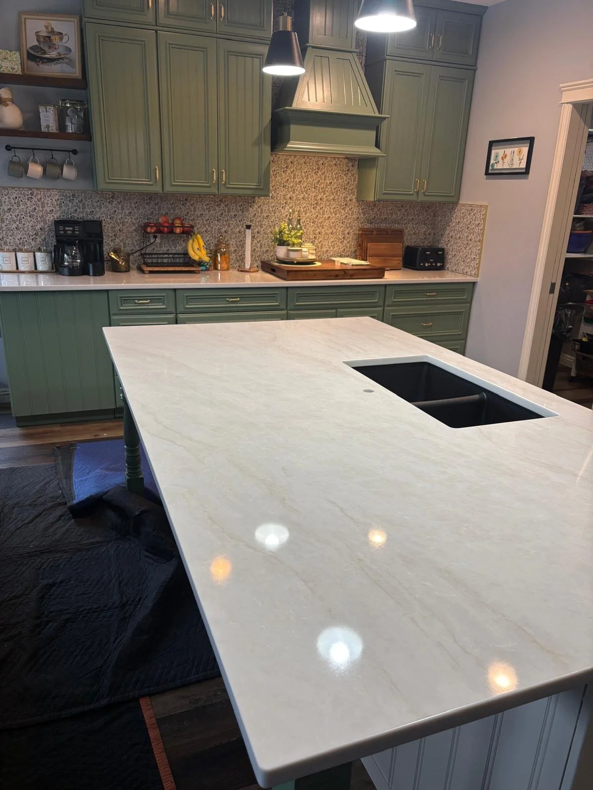 Kitchen with green cabinets, patterned backsplash, and a white marble island with an undermount sink, modern black pendant lights, and various small appliances and decor.