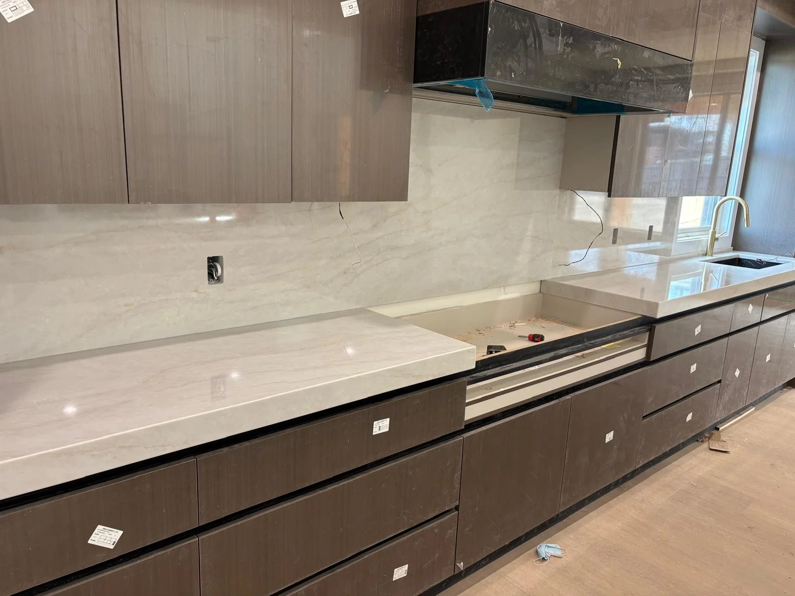 Kitchen with brown cabinets, a white marble countertop and backsplash, a gold faucet, and a window. The kitchen is under construction with tools and wires visible.