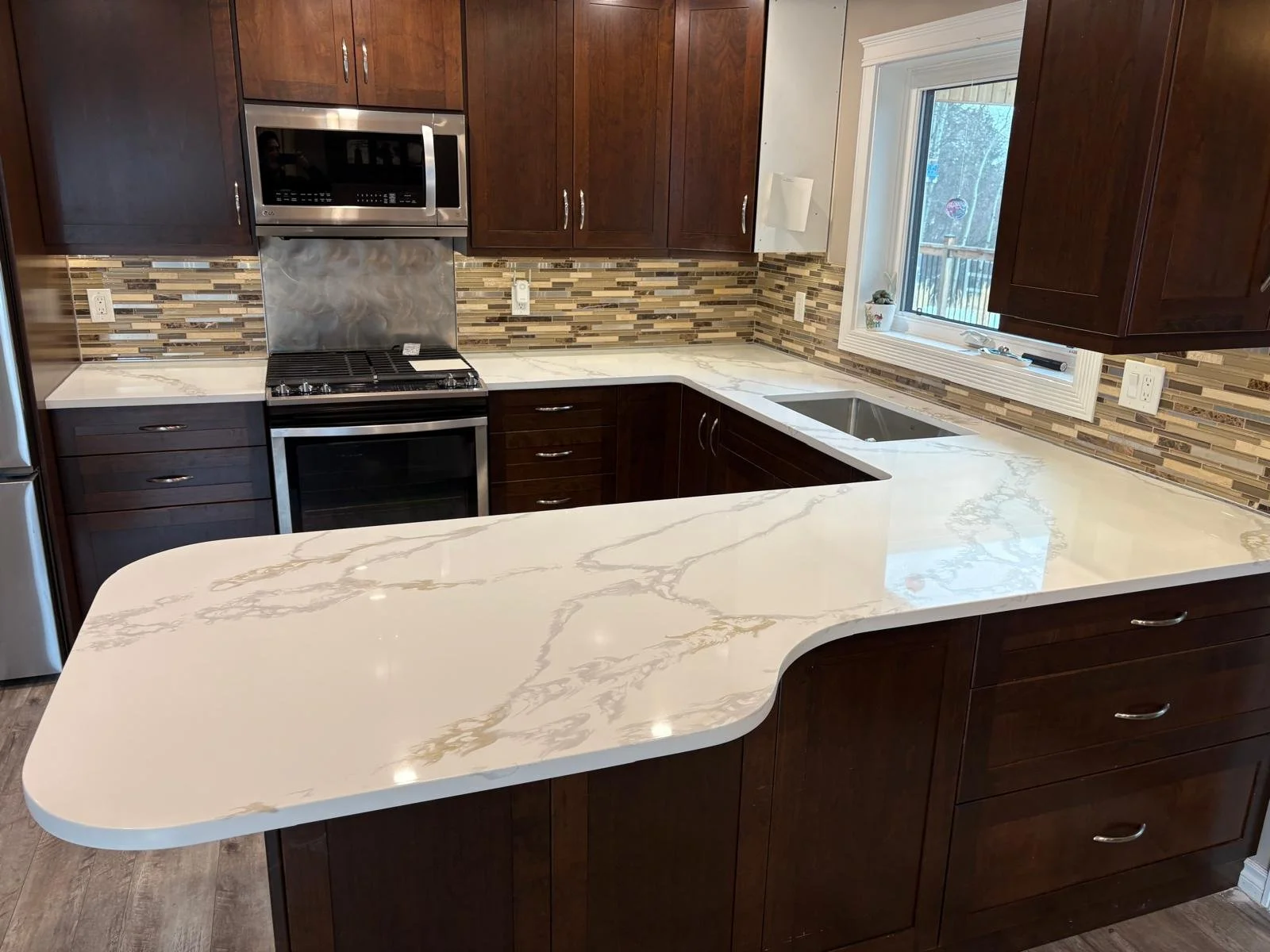 Kitchen with dark wood cabinets, a marble countertop, a window over the sink, a stainless steel microwave and stove, and a modern backsplash with multicolored tiles.