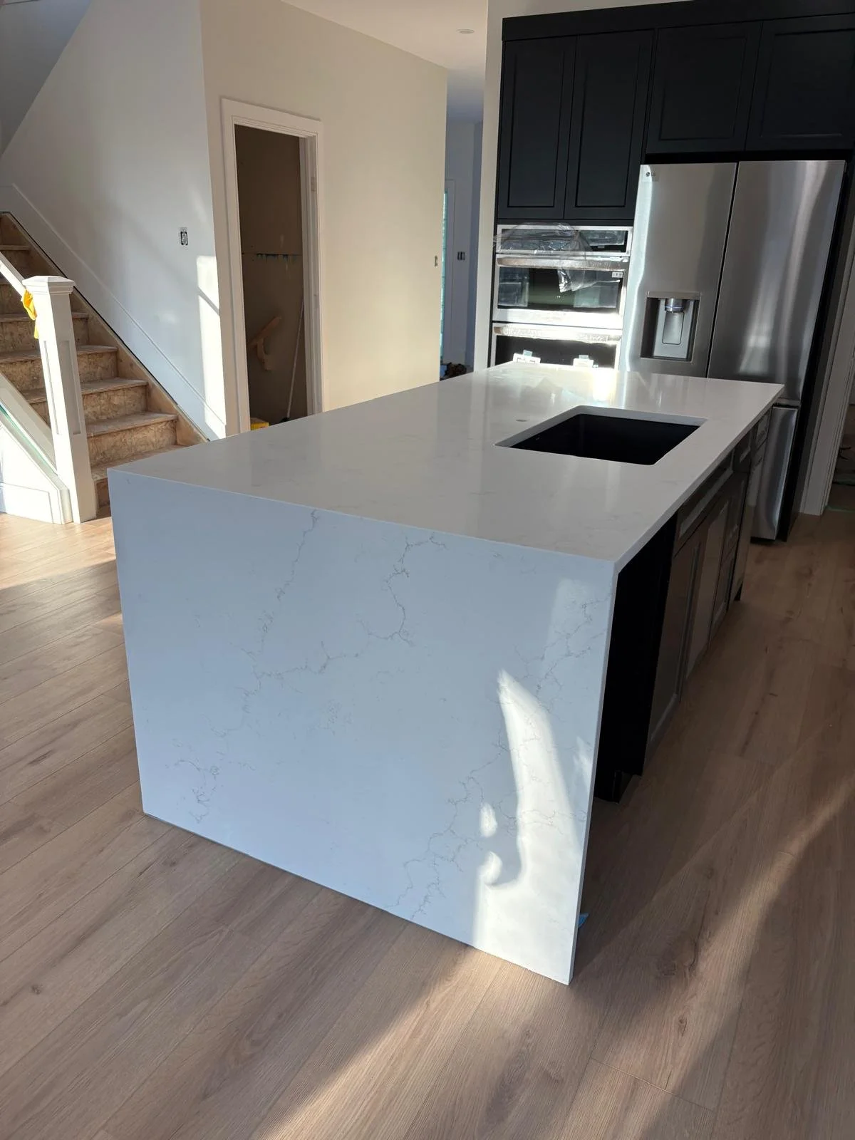 Modern kitchen island with a white marble-like countertop and dark cabinet base, stainless steel refrigerator and built-in oven, stairs and doorway in background.