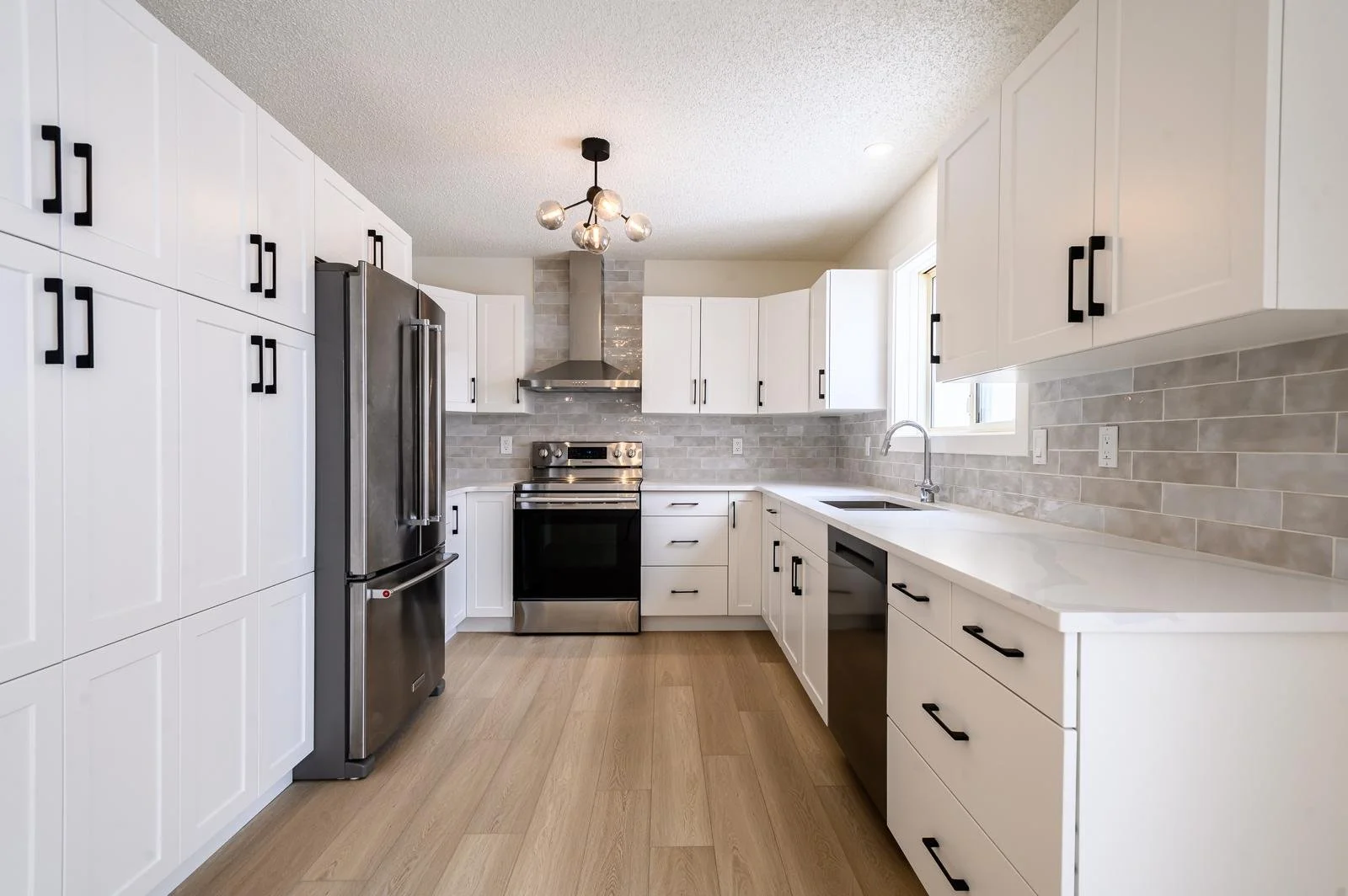 Modern kitchen with white cabinets, stainless steel appliances, a black oven, and beige tile backsplash, with natural light coming through a window.