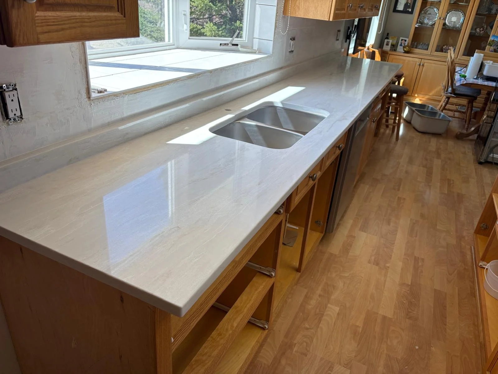 A kitchen countertop with a double sink, wooden cabinets, and a window overlooking trees, with chairs and a table in the background.