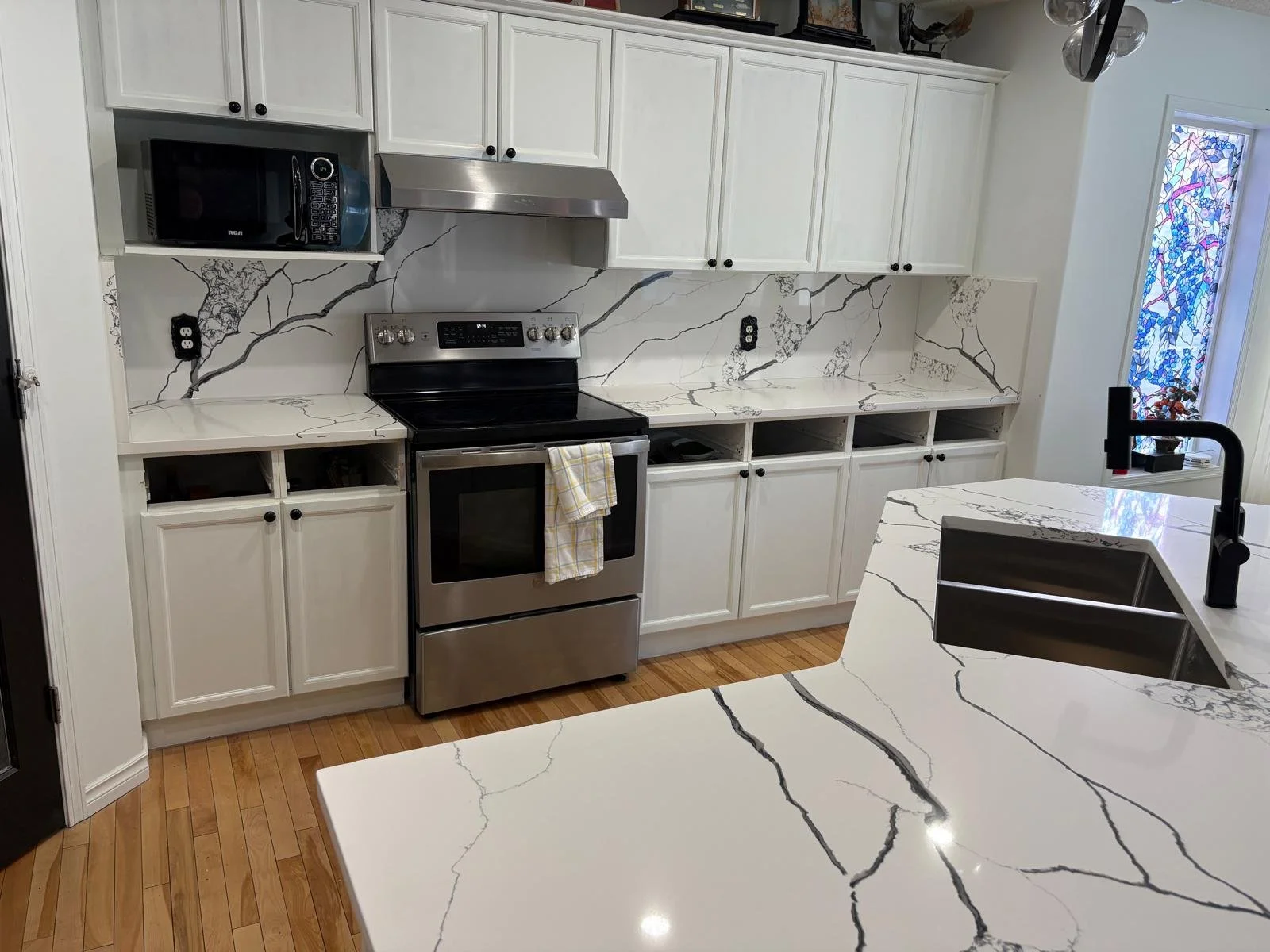 Modern kitchen with white cabinets, black hardware, and black and white marble countertops. Includes stainless steel stove, microwave, and a black kitchen faucet over the sink. Hardwood floor and decorative window with colorful stained glass.