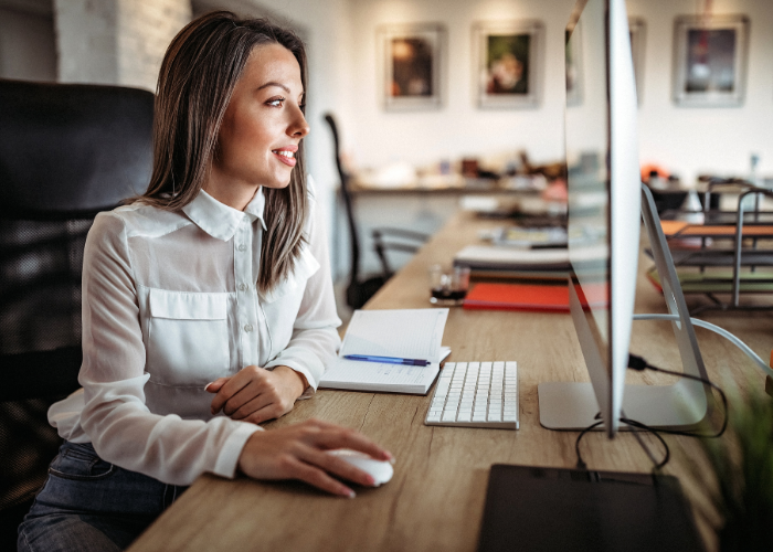 Woman working at a desk with a computer and notepad in an office setting.