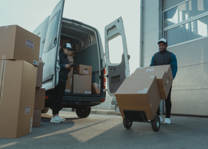 Two people unloading cardboard boxes from a delivery van outside a modern building, with one person pushing a hand truck carrying boxes.