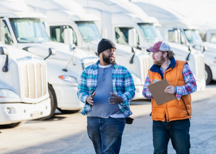 Two truck drivers conversing in a parking lot with multiple semi trucks in the background.