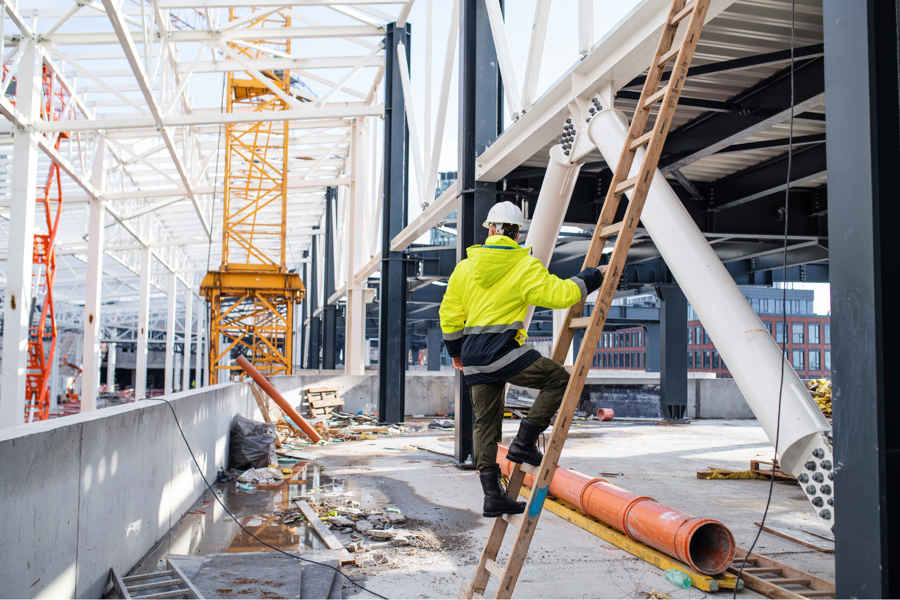 A construction worker in a neon yellow jacket and safety helmet is ascending a wooden ladder on a construction site featuring structural steel framework. The site is cluttered with construction tools, pipes, and debris, with a yellow tower crane and part of a cityscape in the background.
