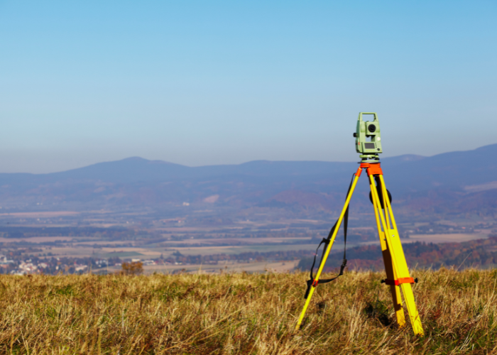 A surveying instrument set up on a yellow tripod in an open field with mountain range in the background under a clear blue sky.