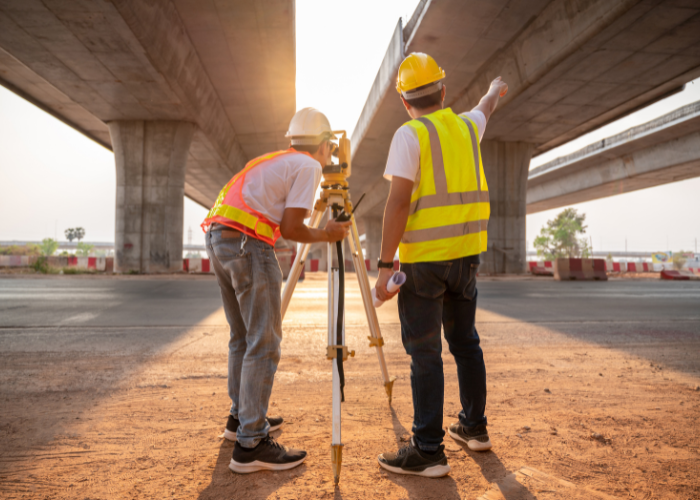 Two construction workers wearing safety vests and helmets working under a bridge, using surveying equipment at a construction site.