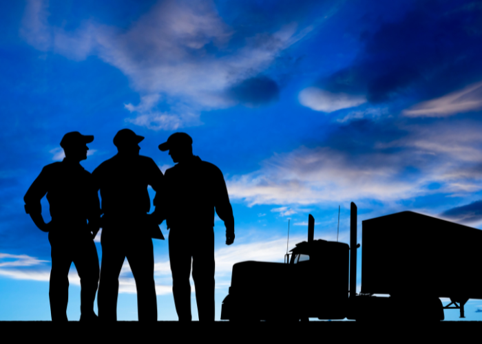 Silhouettes of three people talking near a large truck against a blue sky with clouds.