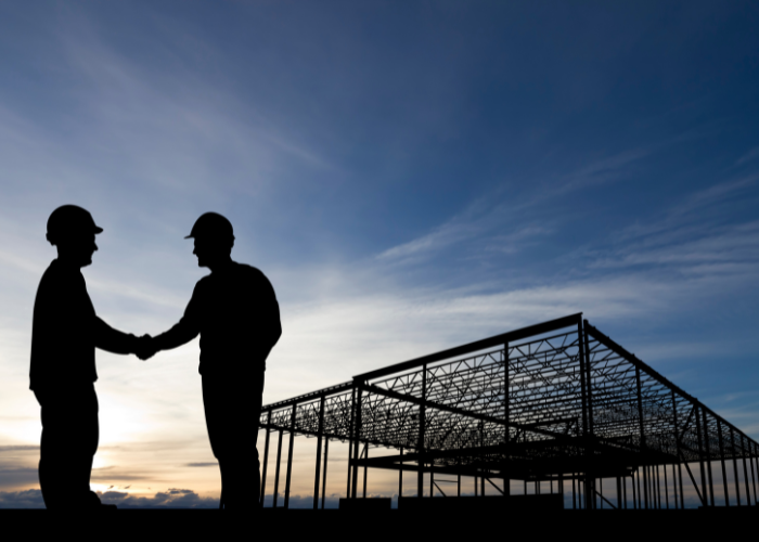 Silhouettes of two construction workers in hard hats shaking hands at sunset, with an unfinished building structure in the background.