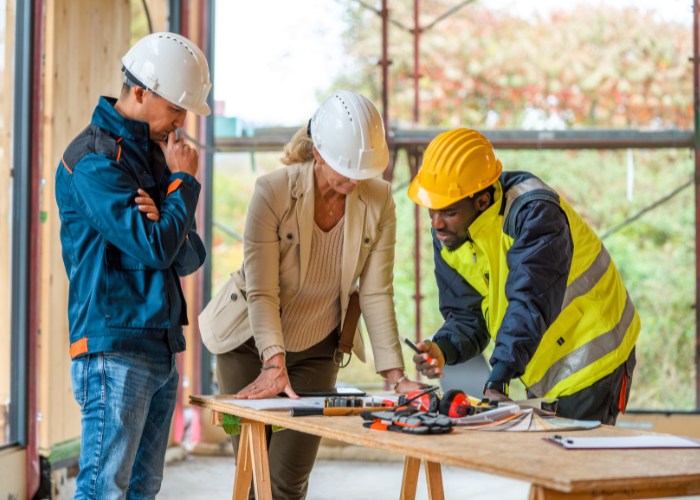 Three construction workers, two men and one woman, wearing safety helmets and work gear, are gathered around a table with tools, discussing plans inside a building under construction with scaffolding and trees visible outside.
