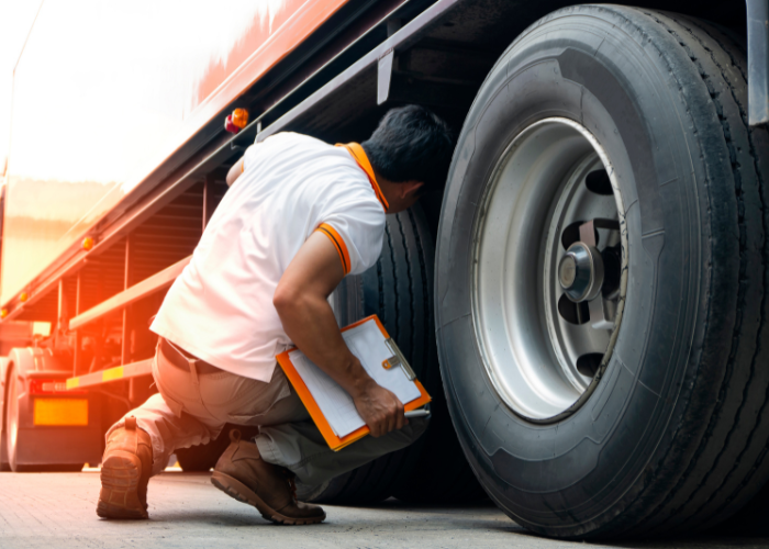 A man inspecting a large truck tire while kneeling on the ground, holding a clipboard.