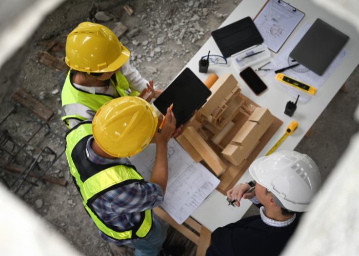Three construction professionals wearing safety helmets and vests examining a wooden architectural model at a construction site, with measuring tools, blueprints, and laptops on the table.