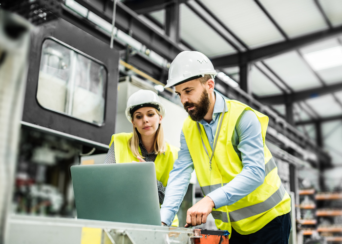 Two engineers wearing safety helmets and reflective vests working with a laptop in an industrial facility