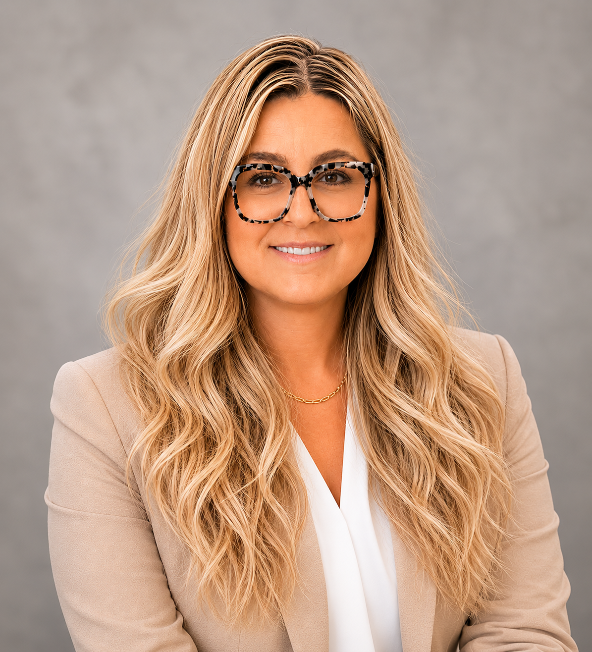 Portrait of a smiling woman with long, wavy blonde hair, wearing large tortoise-shell glasses, a beige blazer, a white blouse, and a gold chain necklace, against a gray background.