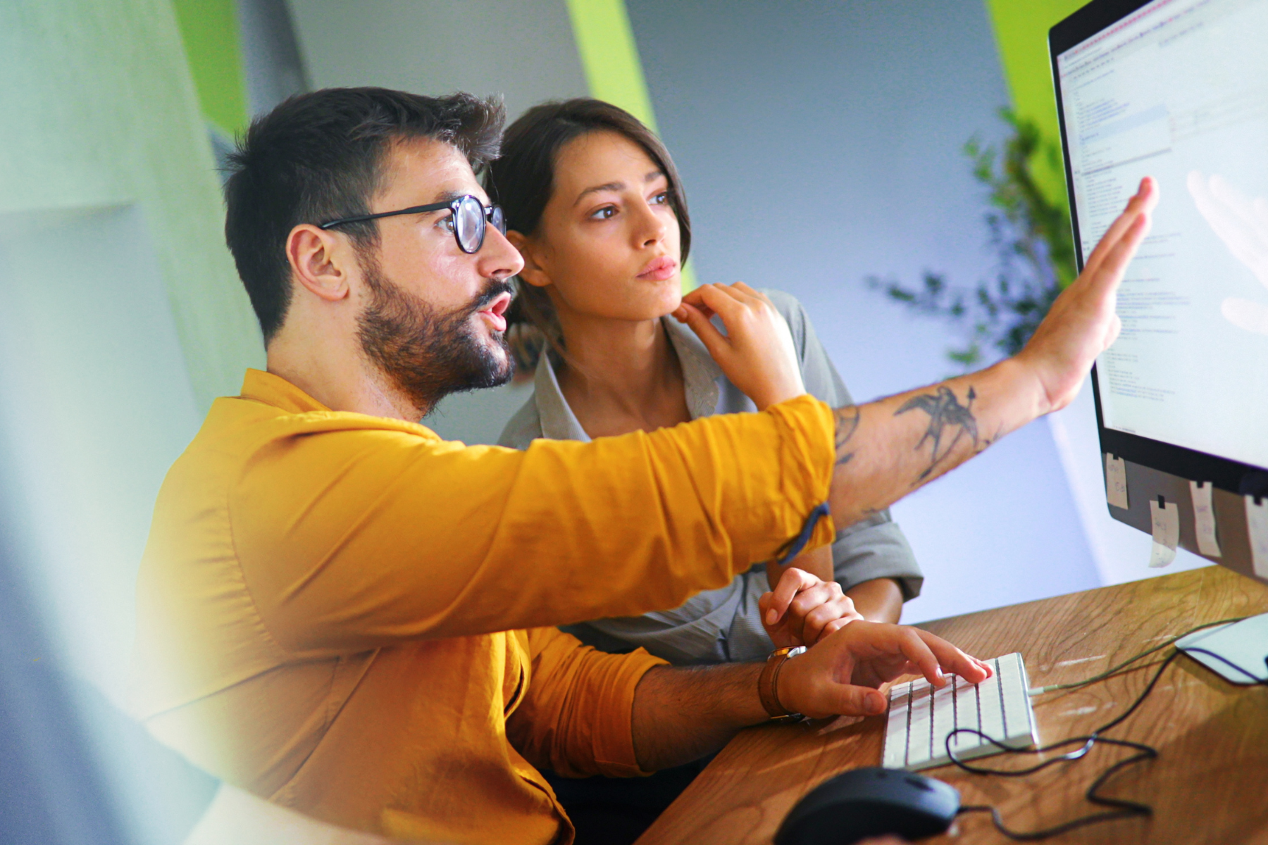Two colleagues work together at a computer, with one pointing at the screen and the other observing, in an office with green and gray walls.