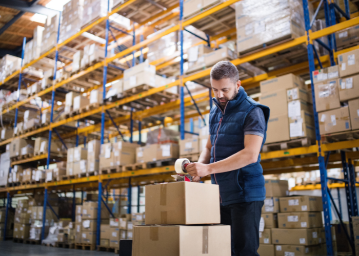 A man in a warehouse is sealing a cardboard box with packing tape.