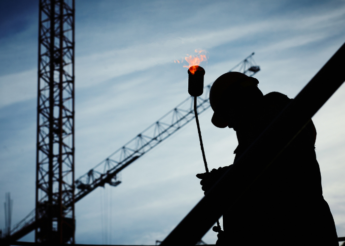 Silhouetted construction worker holding a lit torch with a crane and construction site in the background during dusk or dawn.