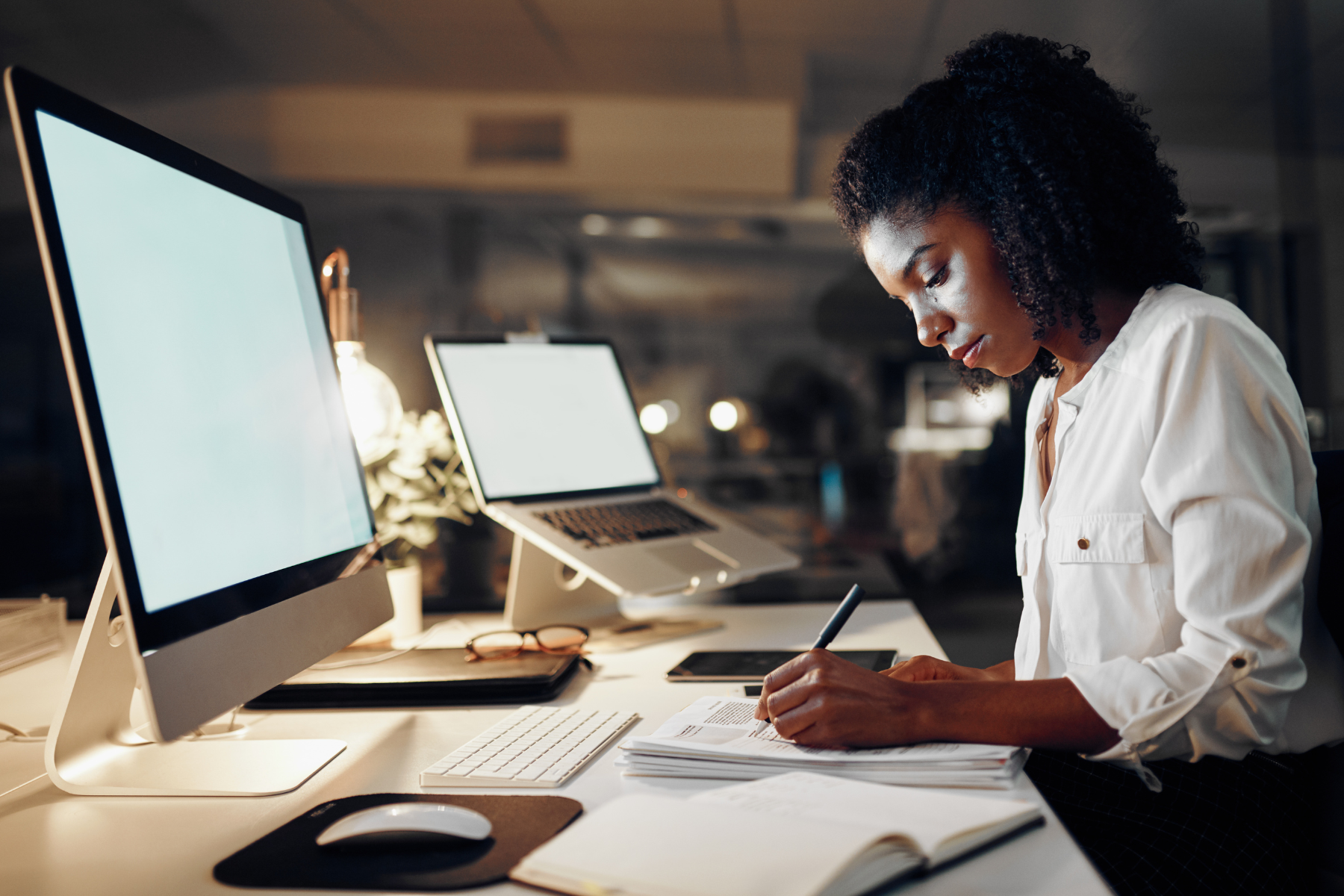 A woman with curly hair wearing a white shirt, working at a desk with multiple computers, notebooks, and a tablet in a modern office setting.
