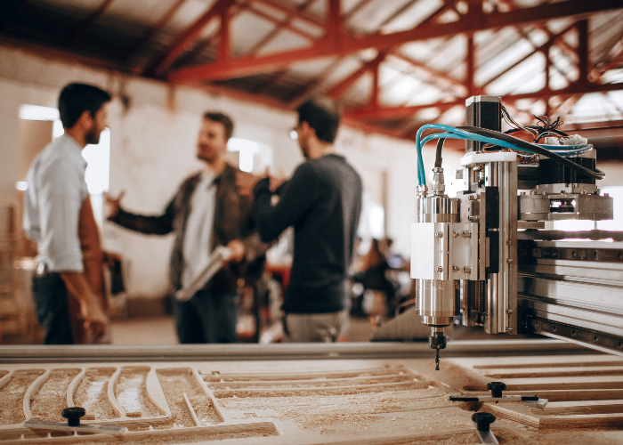 Close-up of a CNC machine or laser cutter at work in a woodworking or manufacturing space with three people in conversation blurred in the background.