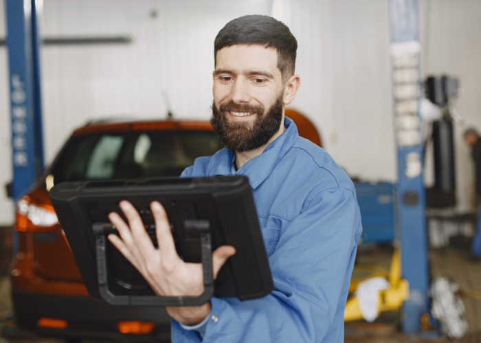 A smiling man in a blue mechanic's shirt looking at a diagnostic tablet inside an automotive workshop, with a car and automotive tools in the background.