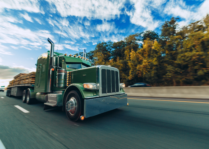 A green semi-truck driving on a highway with trees and a blue sky with clouds in the background.