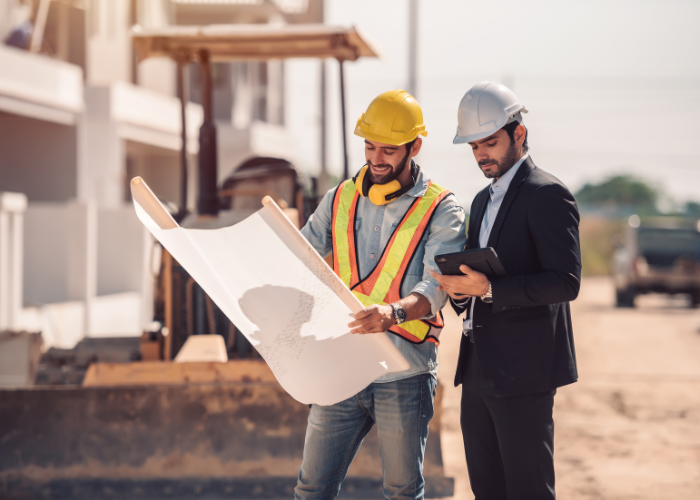 Two construction professionals, one in a yellow safety helmet and vest, the other in a suit and white safety helmet, review blueprints at a construction site.