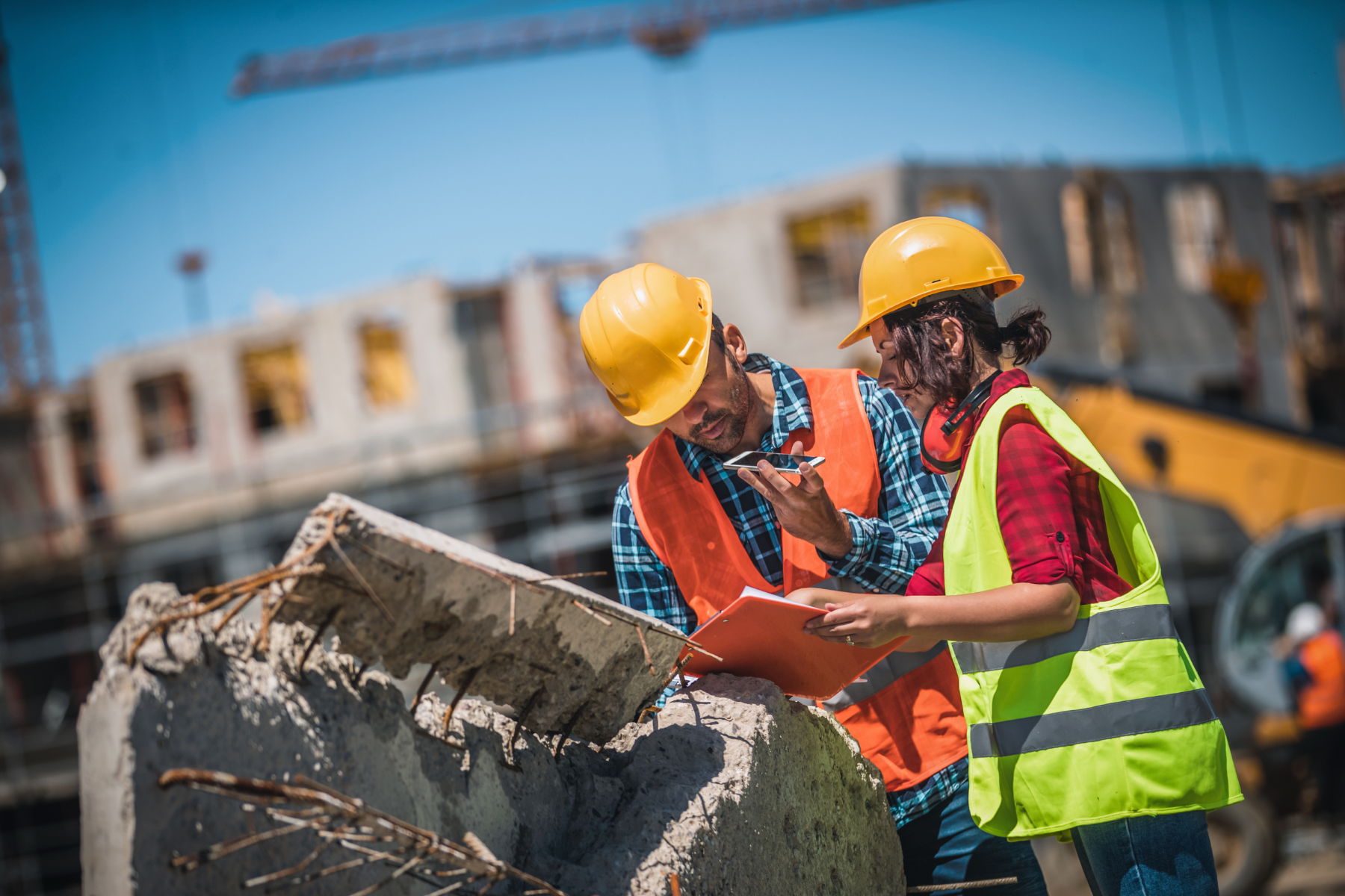 Two construction workers wearing yellow hard hats, safety vests, and checkered shirts at a construction site, reviewing documents and discussing a concrete structure with exposed rebar.