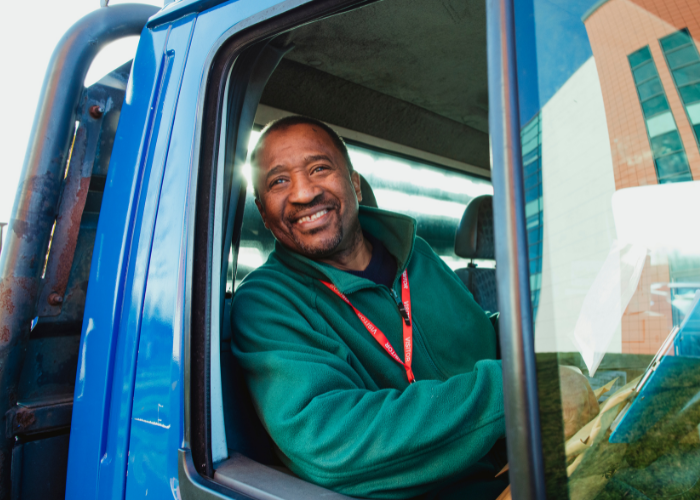 A man smiling and sitting in the driver's seat of a blue truck, outside a modern building.