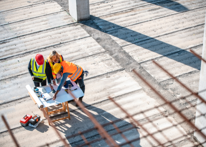 Construction workers with safety helmets and vests working on a blueprint at a construction site under sunlight.