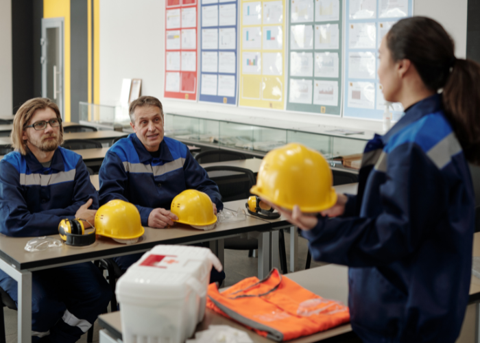 An instructive safety training session in a classroom with three men wearing blue uniforms, sitting at desks with yellow safety helmets and headphones, listening to a woman in a blue uniform demonstrating safety gear with a yellow helmet in her hands. The classroom has charts and posters on the wall.