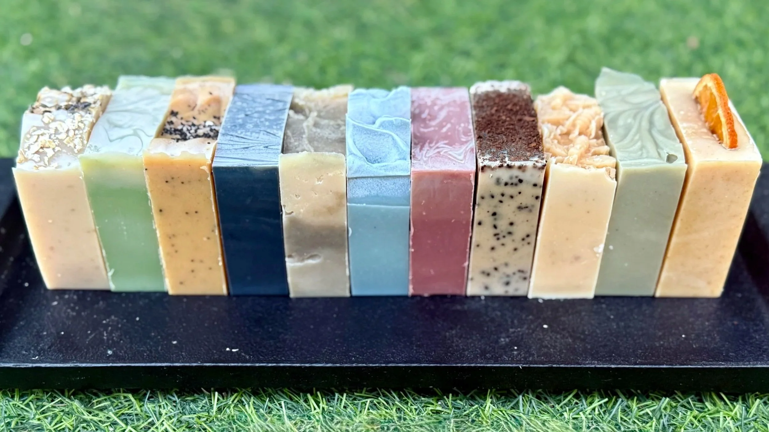 Line of assorted handmade soap bars with various colors and textures, placed on a black surface outdoors.