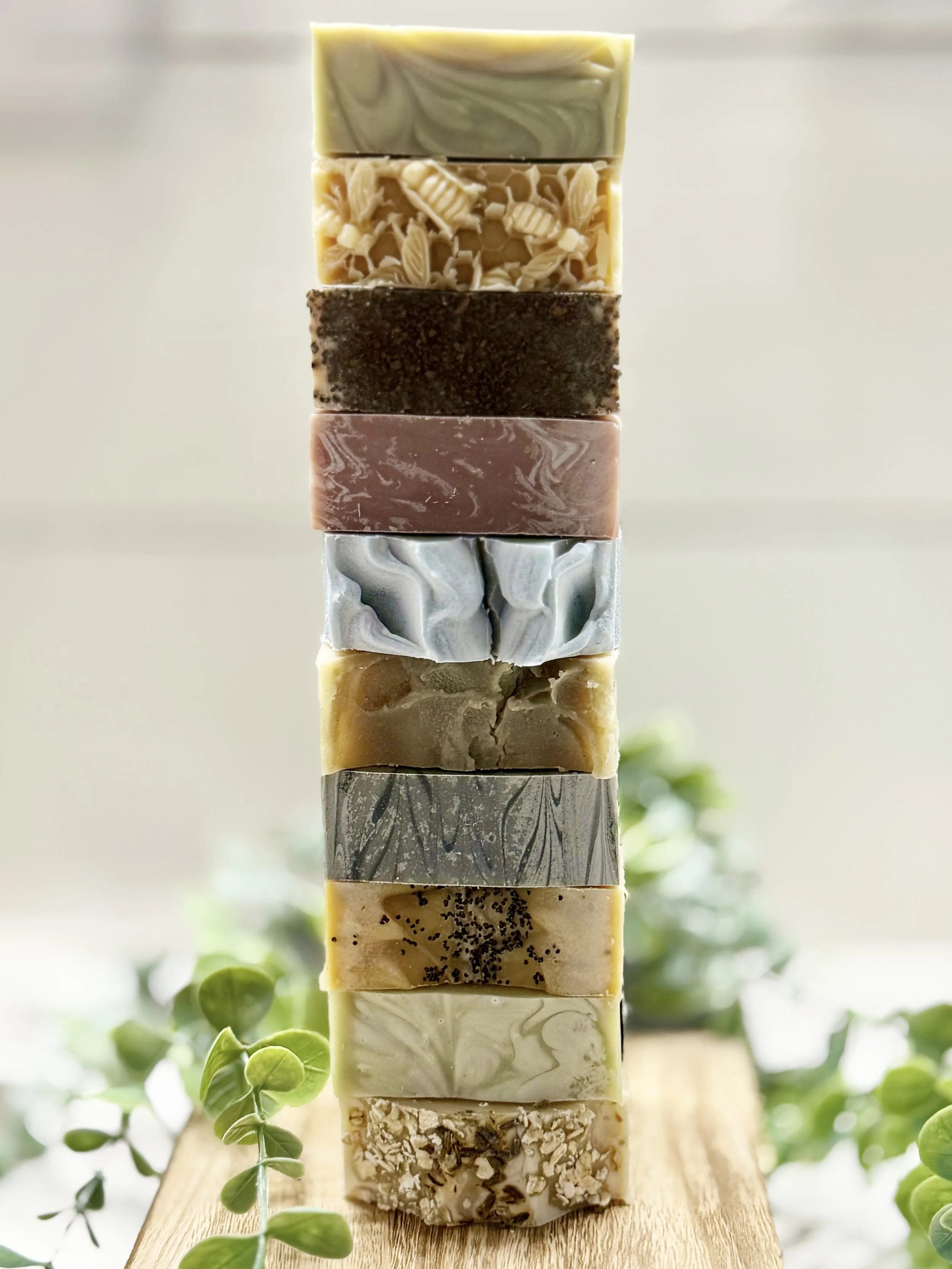 Stack of nine handmade soap bars with various colors and patterns, arranged vertically on a wooden surface with green foliage in the background.