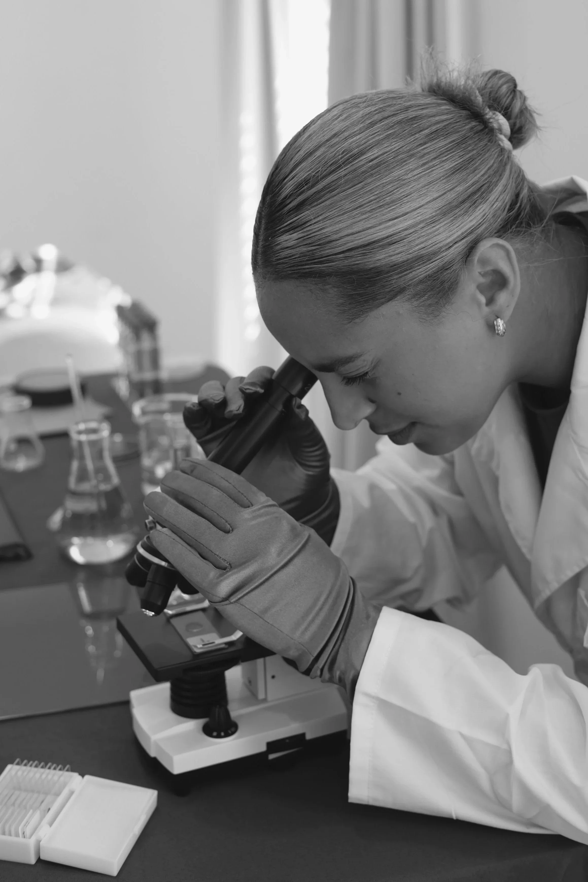 A woman with blonde hair in a bun, wearing earrings and a lab coat, looking into a microscope in a science laboratory with test tubes and scientific equipment on the table.