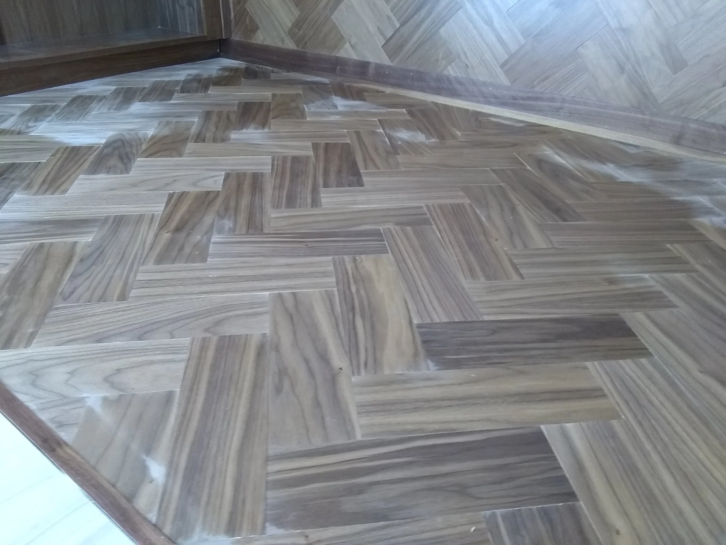 Close-up image of a wooden parquet floor with a pattern of rectangular tiles in varying shades of brown, showing a corner where the floor meets the wall and a wooden edge.