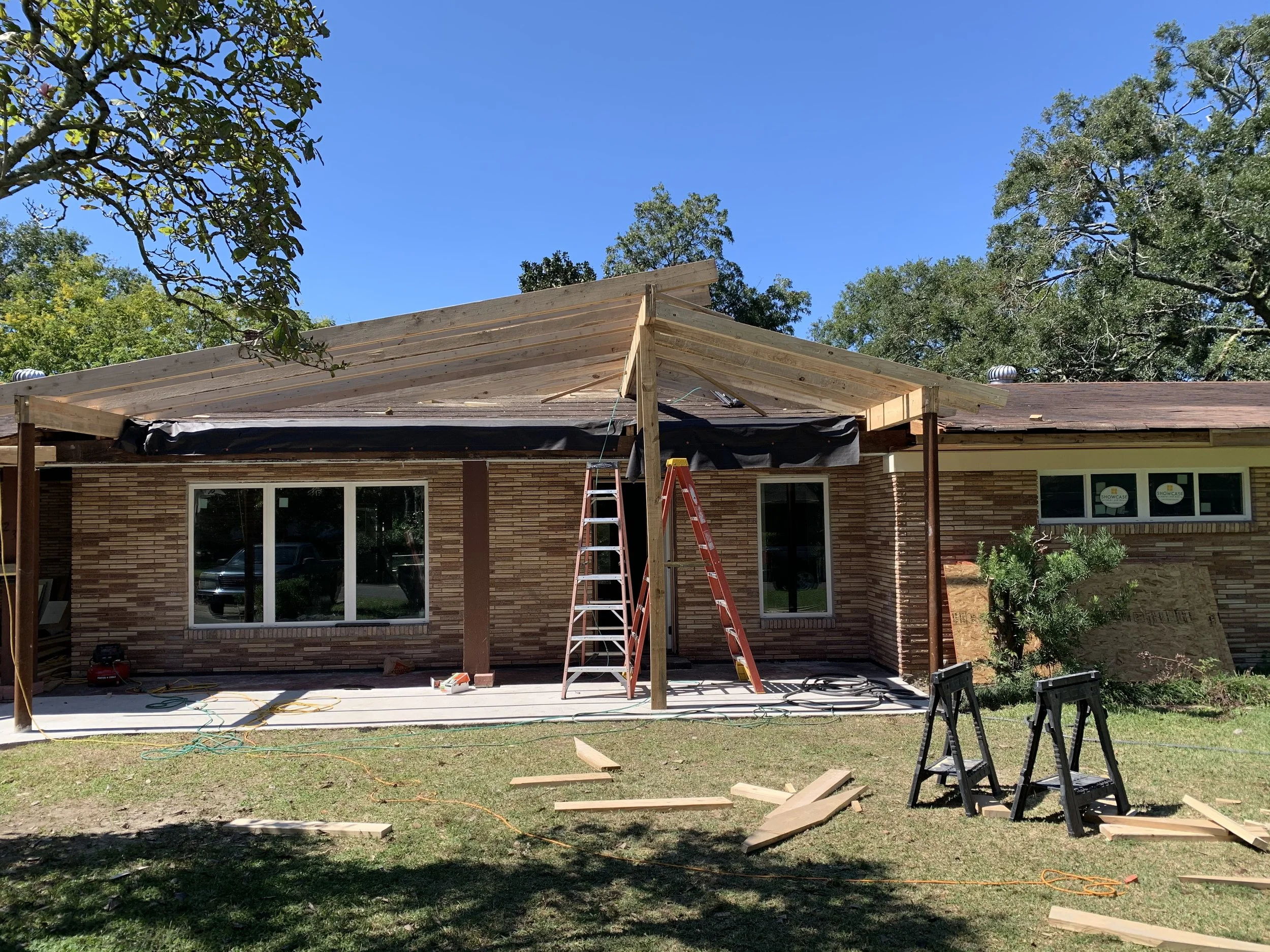 Construction site of a house extension with wooden framing and roofing work in progress. Tools and construction materials are present on the ground, and a ladder is leaning against the framing.