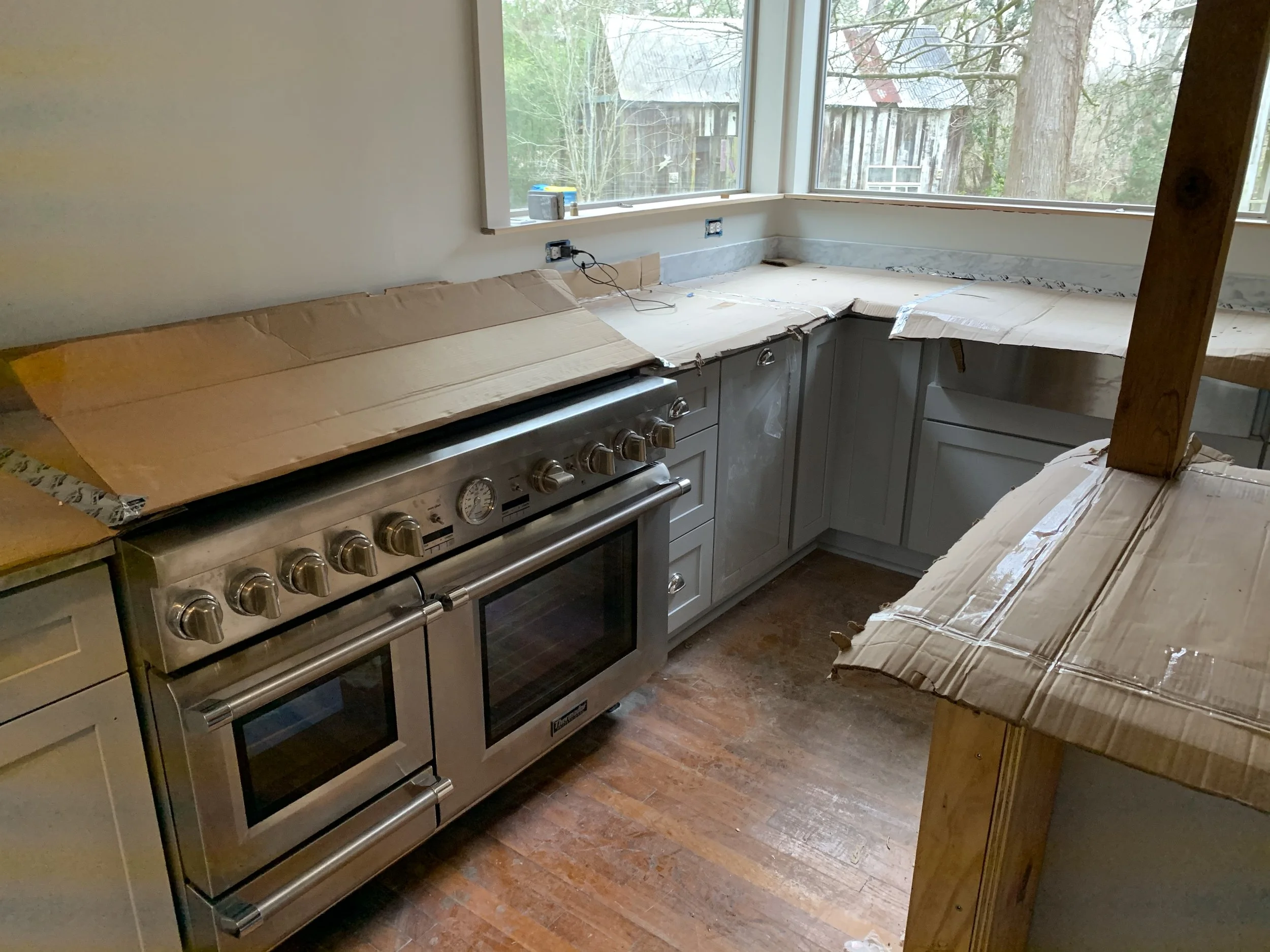 Kitchen with a stainless steel stove, white cabinets, and a large window showing an outdoor yard with trees and a shed. Cardboard covers parts of the countertop and a wooden armrest is visible.