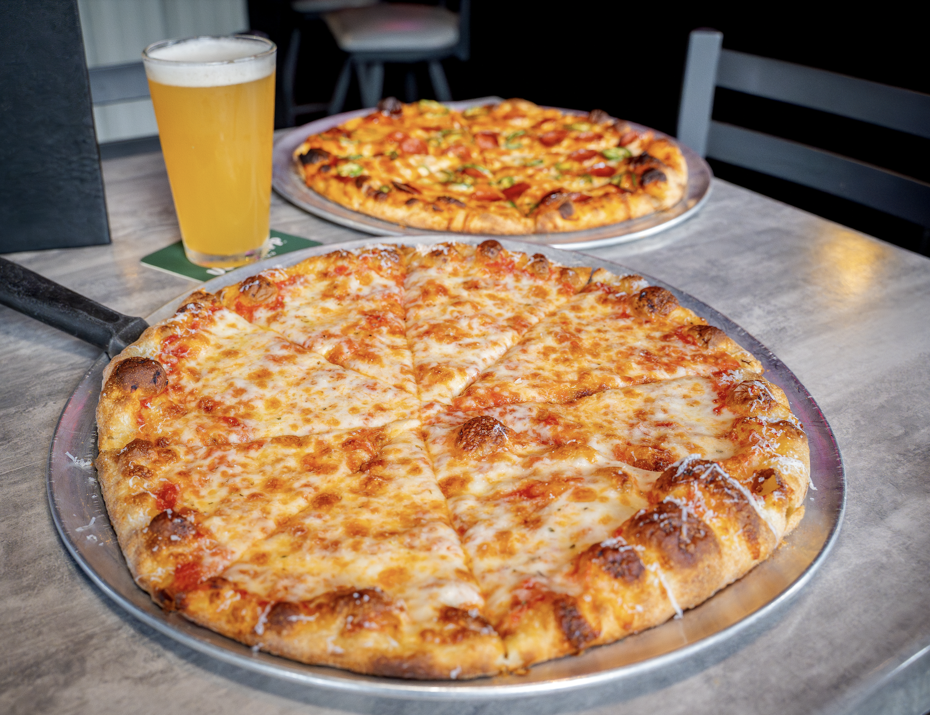 A cheese pizza on a metal tray with another pizza in the background and a glass of beer on a table.