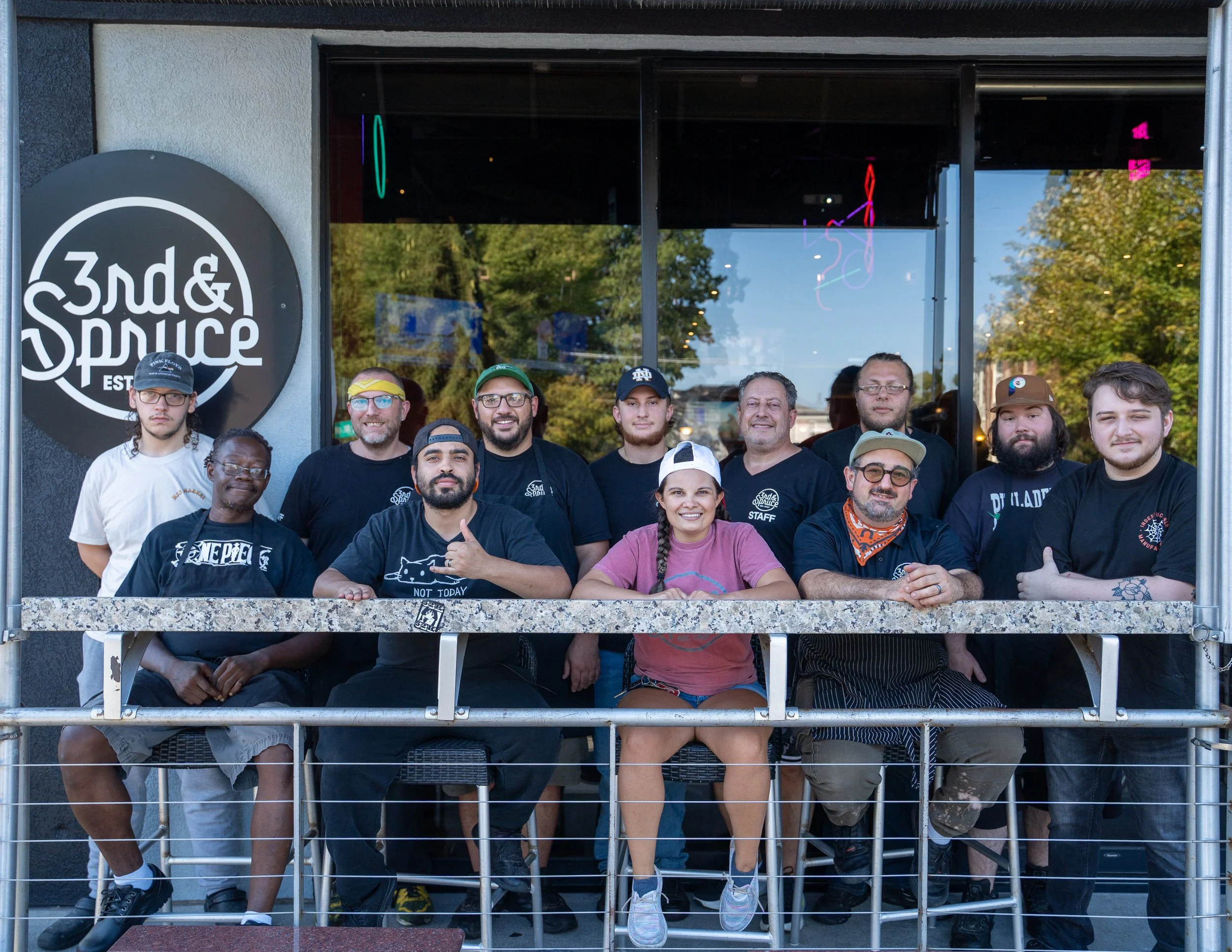 A group of employees smiling and posing in front of the storefront with a sign that reads '3rd & Spruce'. 
