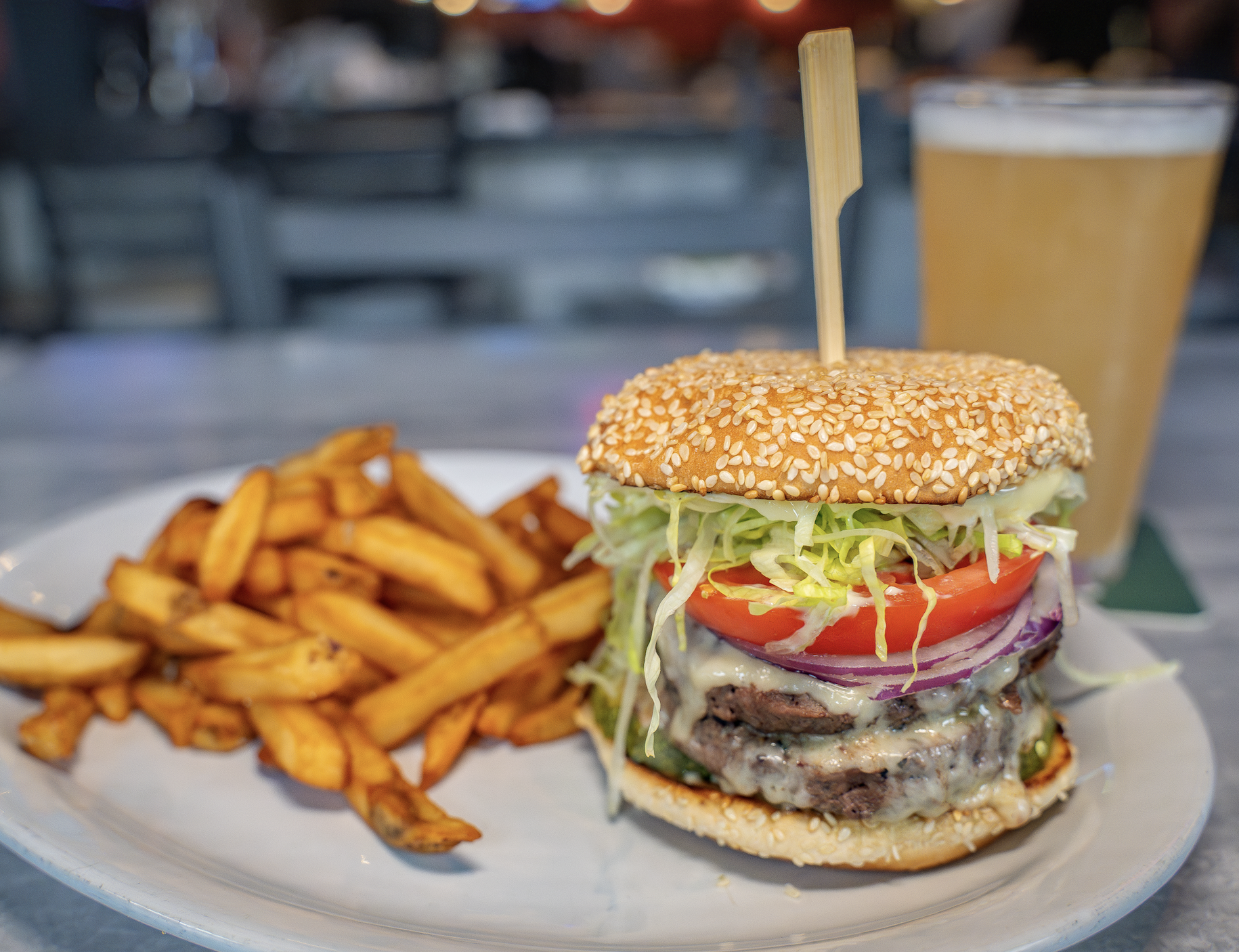 A plate with a cheeseburger topped with lettuce, tomato, onions, and a sesame seed bun, served with a side of French fries. In the background, there is a pint of beer.
