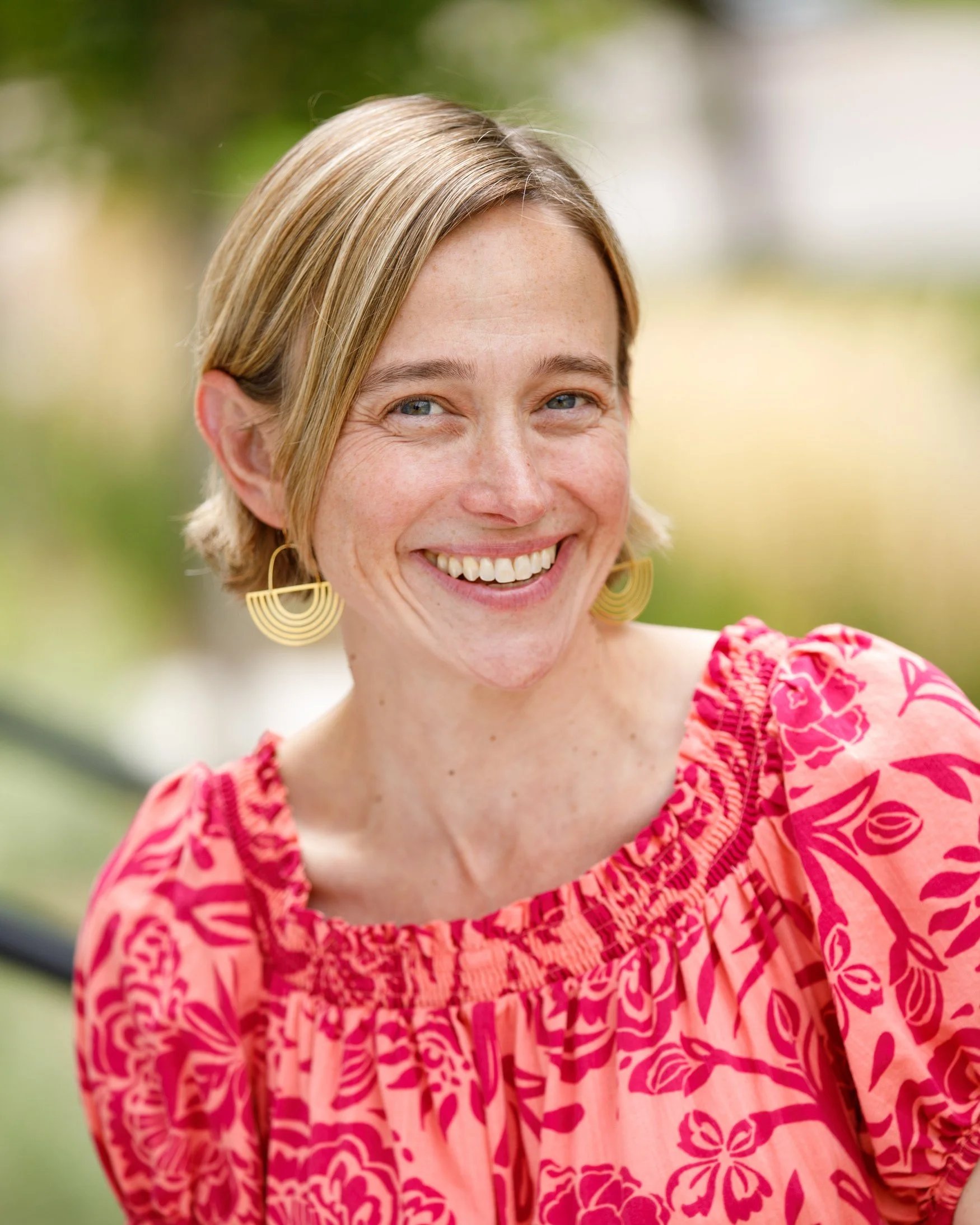 Close-up of a smiling woman with short blonde hair, wearing large yellow earrings and a pink floral dress, outdoors with blurred green background.